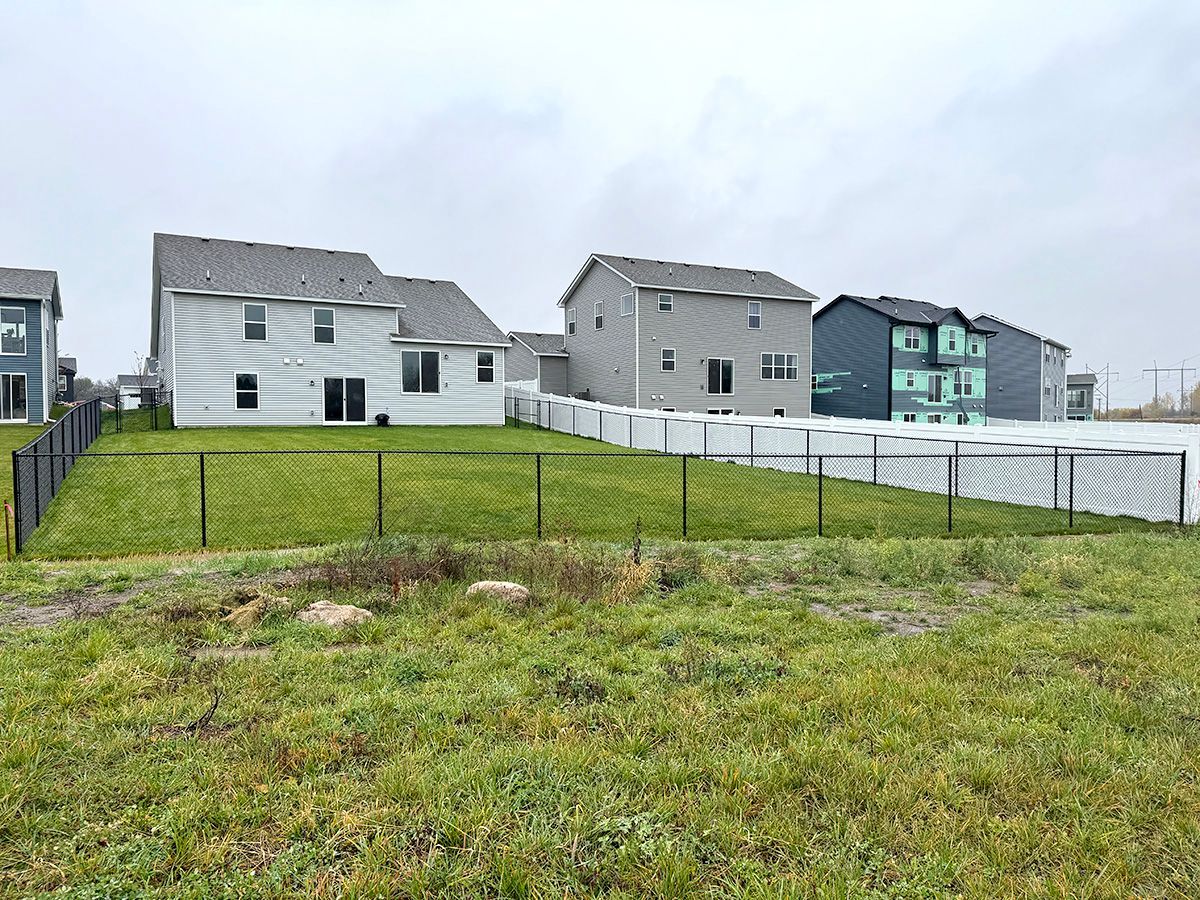A row of suburban houses with fenced-in backyards viewed from a grassy, undeveloped lot under a cloudy sky.
