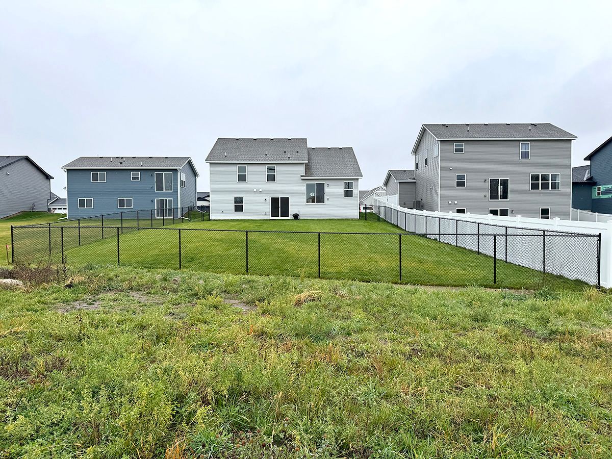 A row of residential houses with fenced-in backyards overlooking an open, grassy field under a cloudy sky.