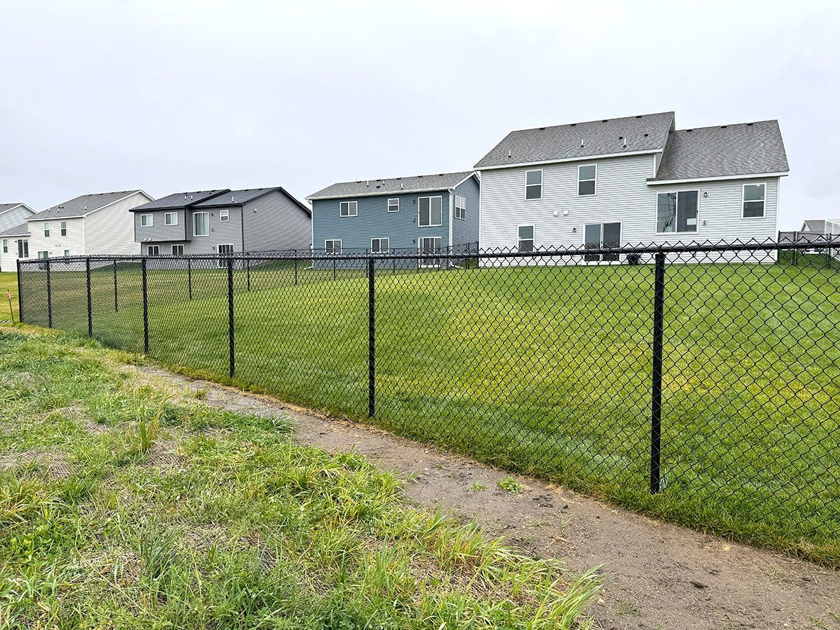 A black chain-link fence lines a grassy backyard behind a row of suburban houses under an overcast sky.