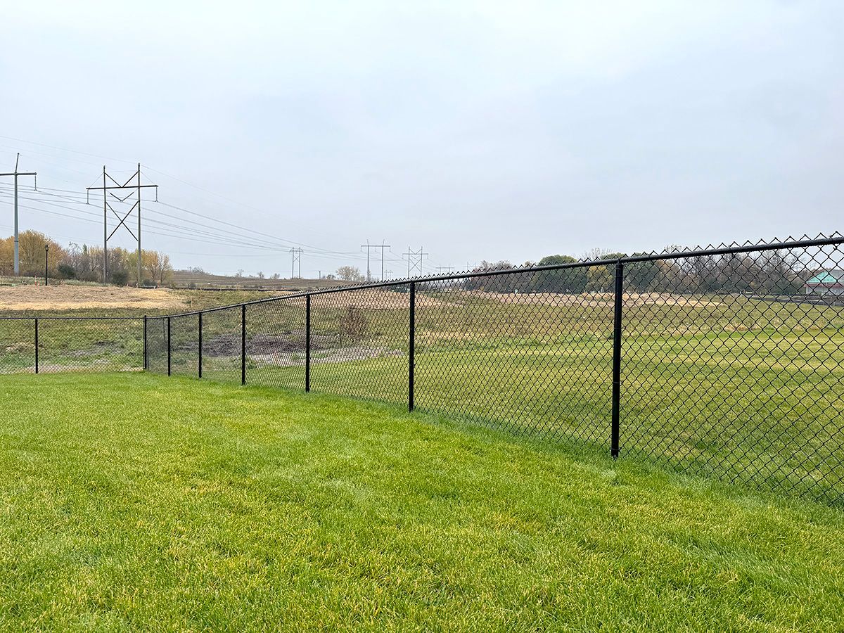 A black chain-link fence runs along a green grassy lawn under a cloudy sky, with power lines in the distance.