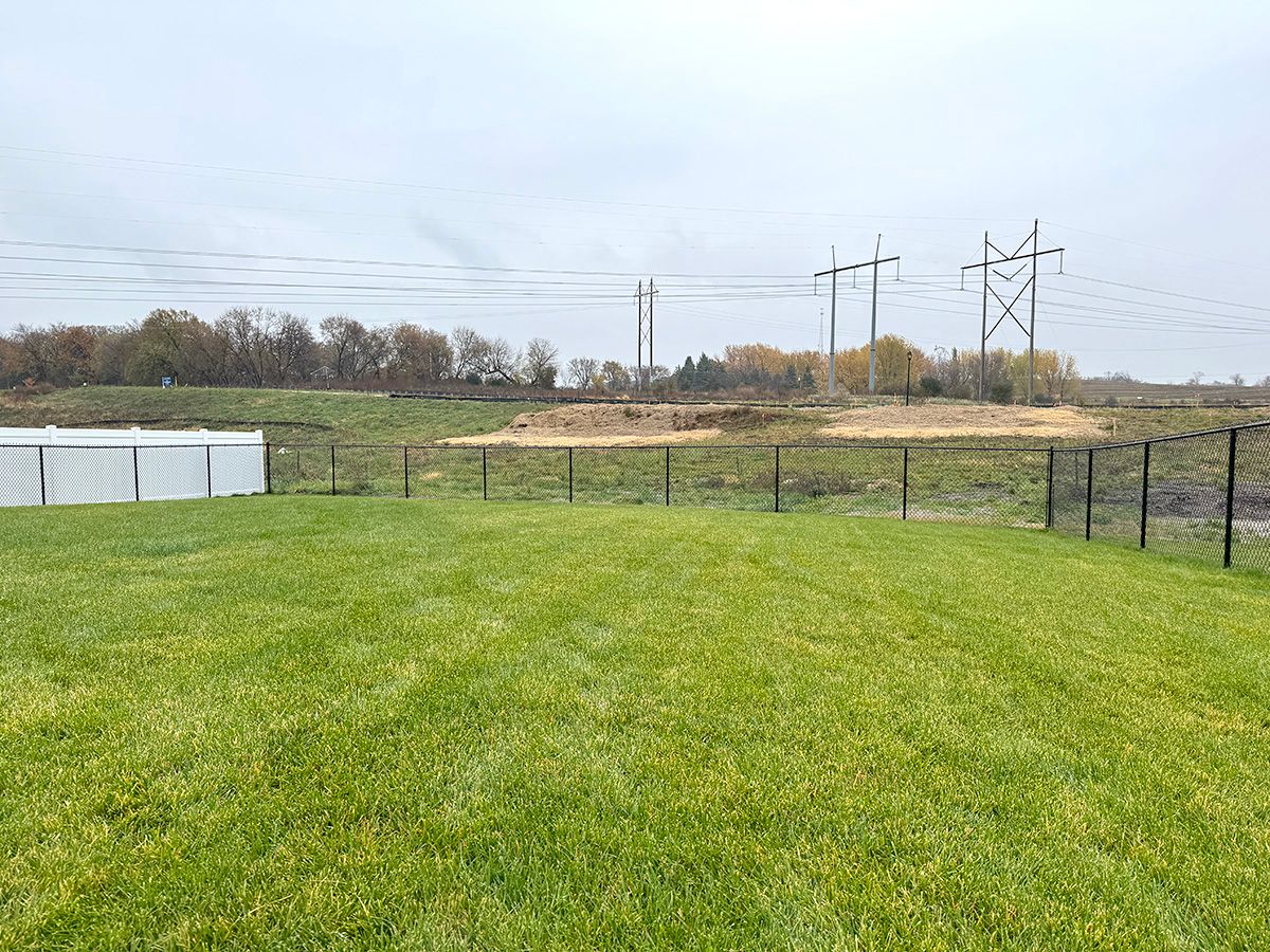 A grassy backyard with a white fence, metal chain-link fencing, and power lines against a cloudy sky.