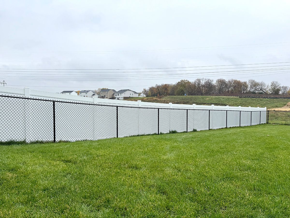 A white chain-link fence with privacy slats runs across a green, grassy yard under a cloudy sky.