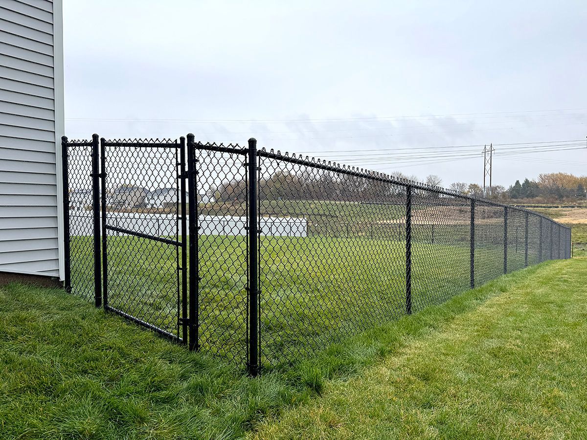 A black chain-link fence with a closed gate attached to the side of a house, bordering a grassy yard.