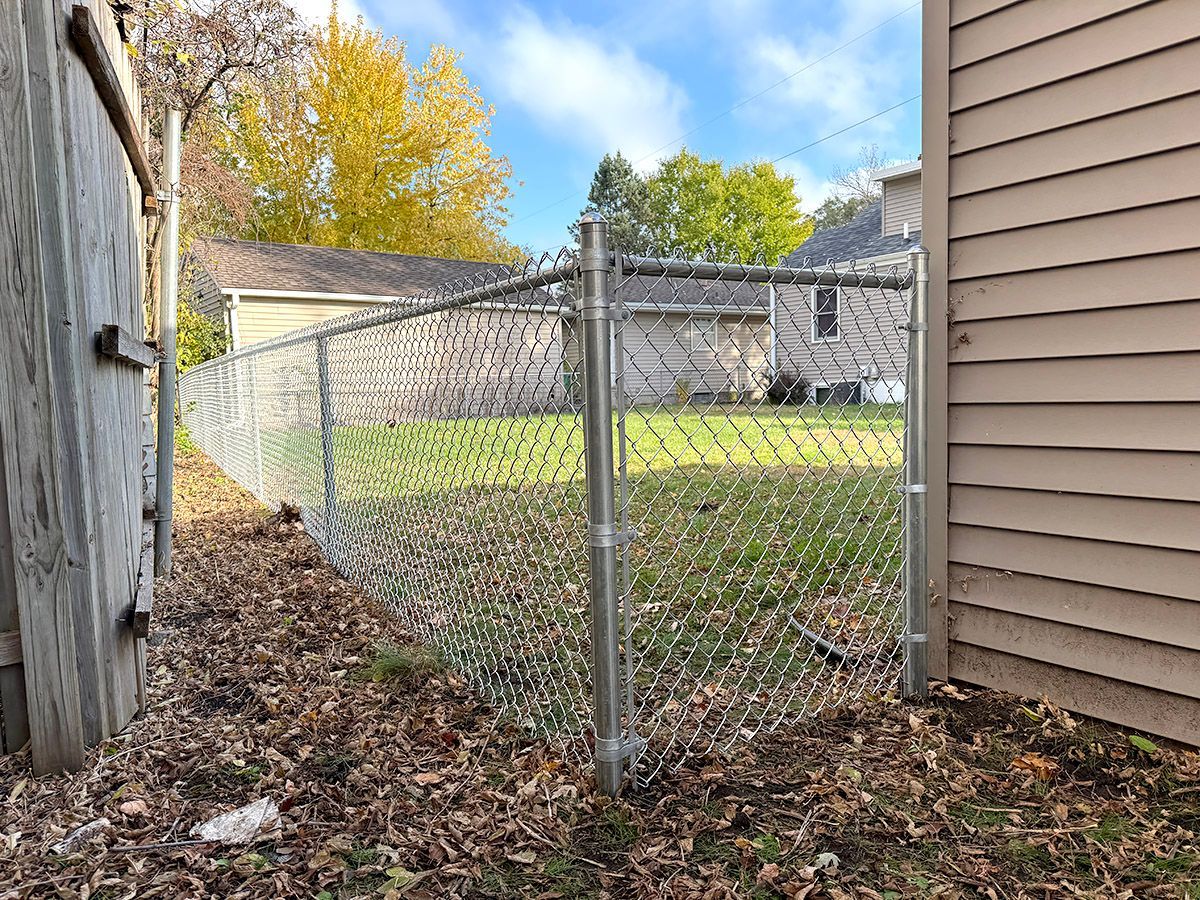 A chain-link fence section corner connects to the beige siding of a house, overlooking a grassy yard with autumn trees.