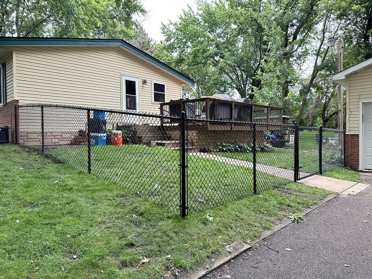 A tan residential house with a wooden deck and a black chain-link fence in a grassy backyard.