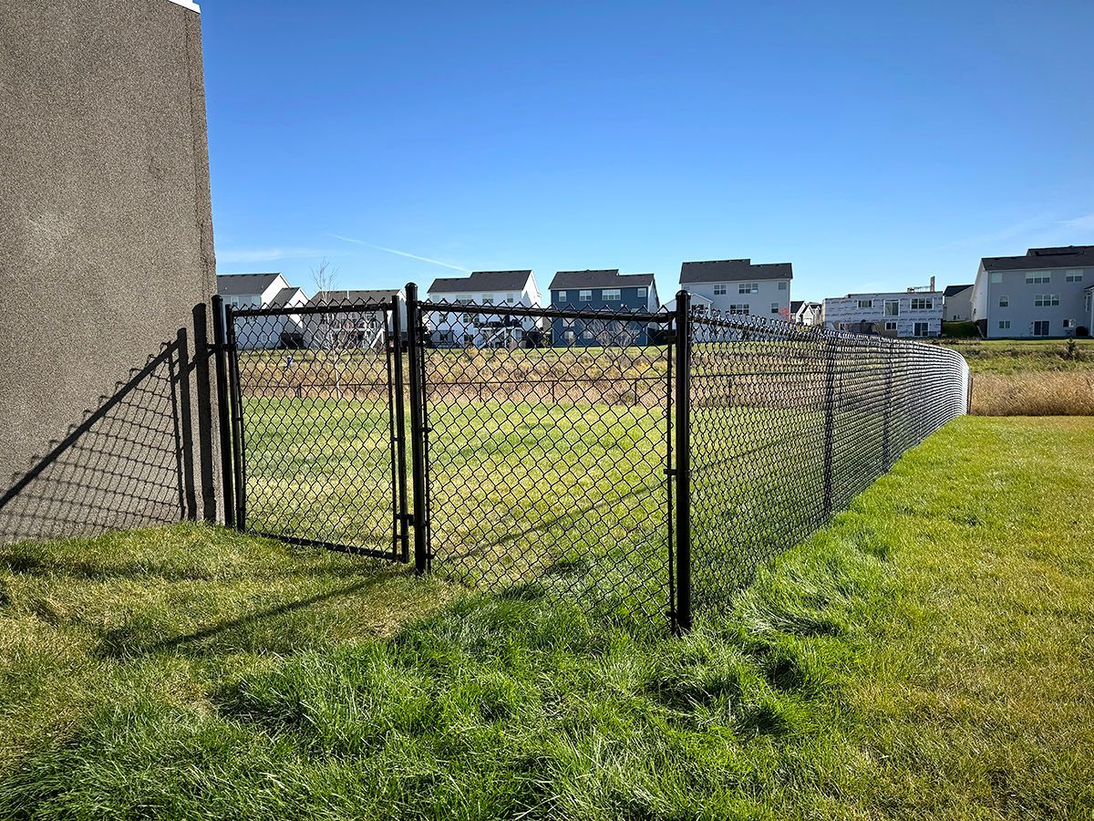 A black chain-link fence with a closed gate attached to a tan building on a sunny day with houses in the background.