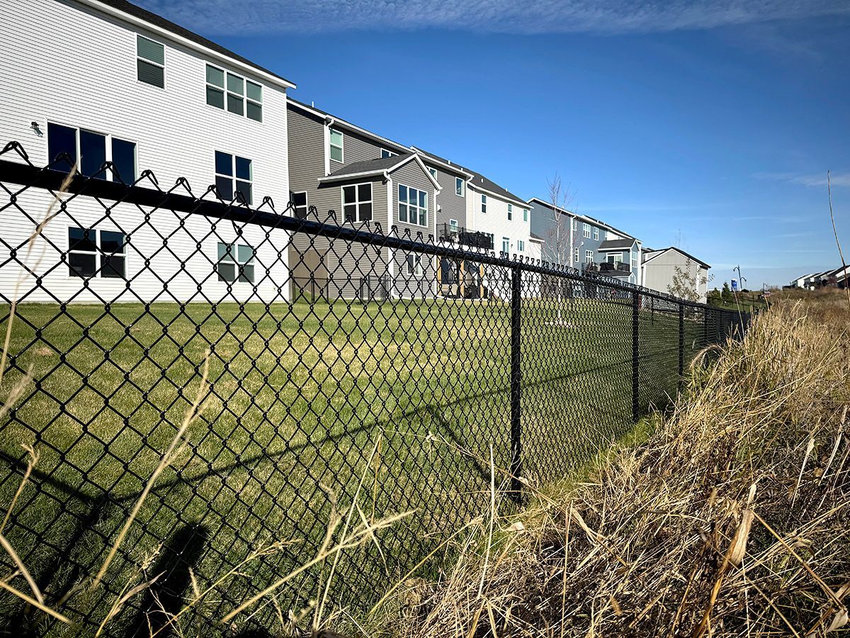 A black chain-link fence lines a grassy slope behind several multi-story suburban houses under a bright blue sky.