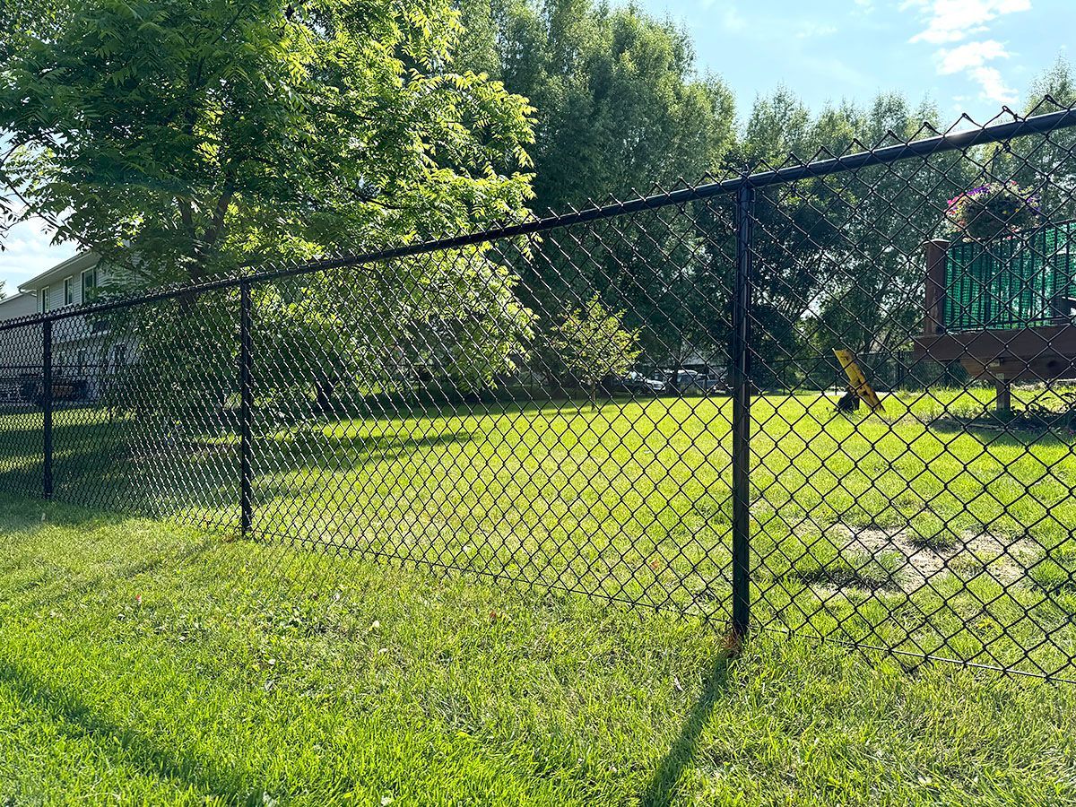 A black chain-link fence encloses a green grassy lawn with trees and a wooden play structure in the background.