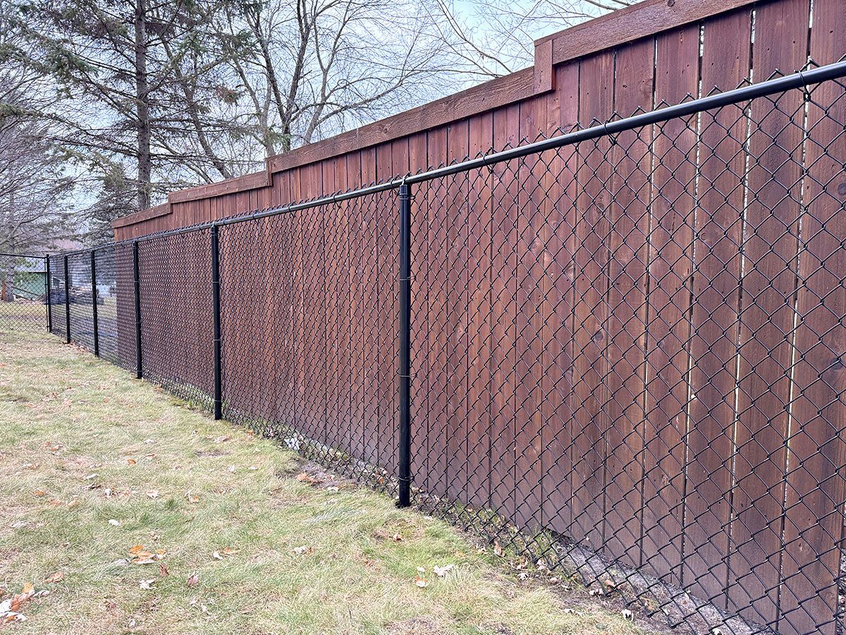 A black chain-link fence stands in front of a weathered, dark wood privacy fence in a grassy outdoor area.