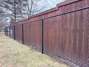 A black chain-link fence runs in front of a wooden privacy fence in a grassy area with trees in the background.