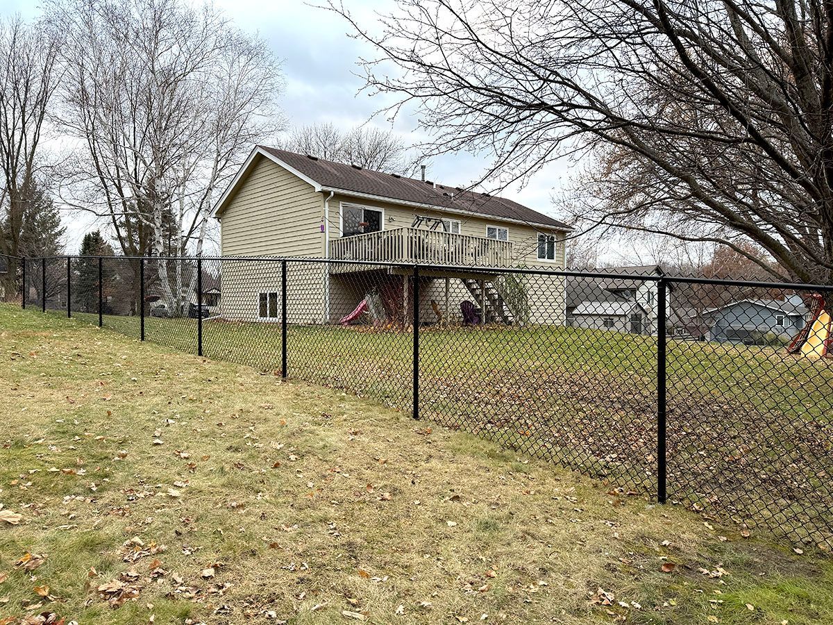 A light-colored house with a raised wooden deck stands behind a black chain-link fence in a grassy yard under a cloudy sky.