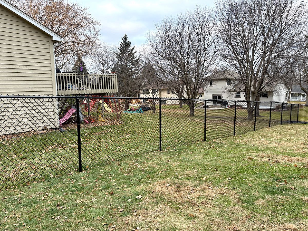 A black chain-link fence divides a backyard, with a house and deck on the left and trees in the distance.