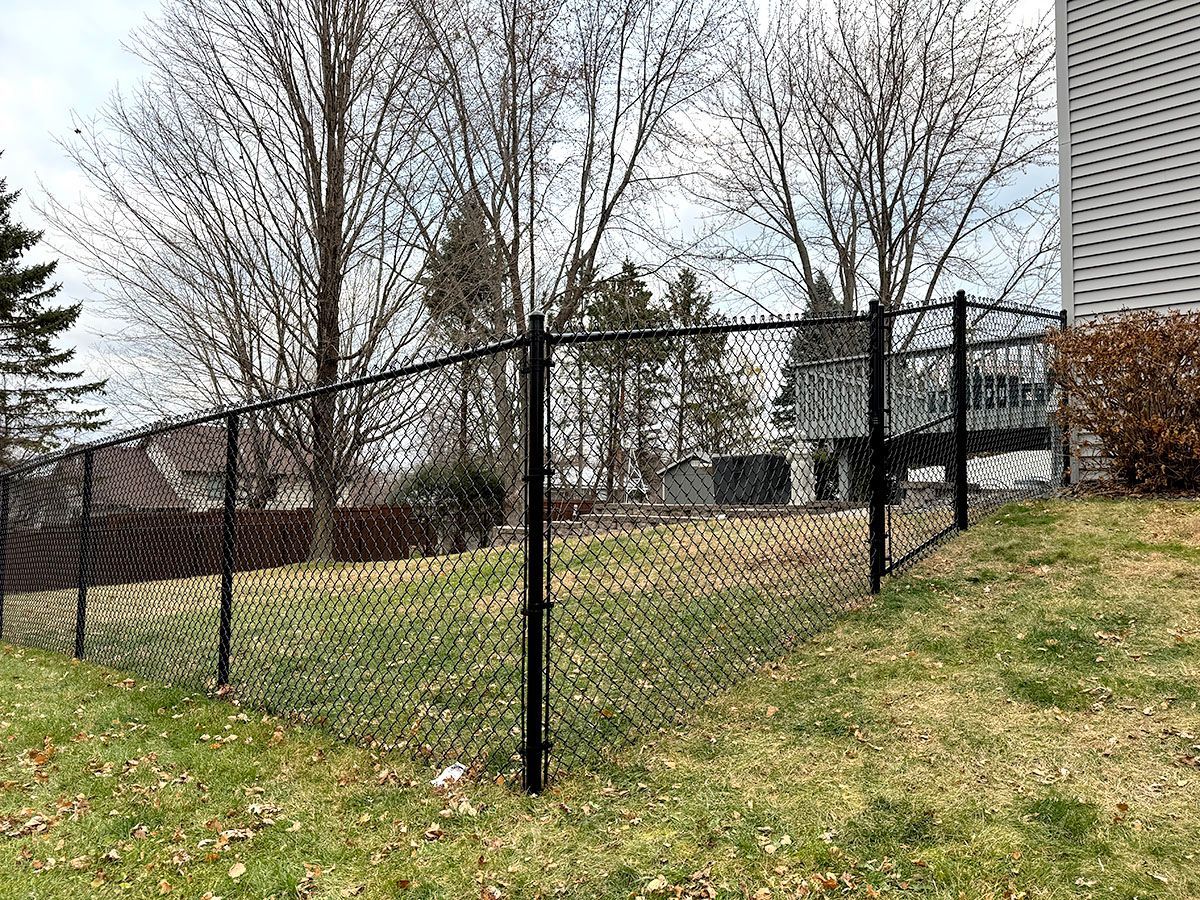 A black chain-link fence lines a grassy backyard next to a house with gray siding on an overcast day.