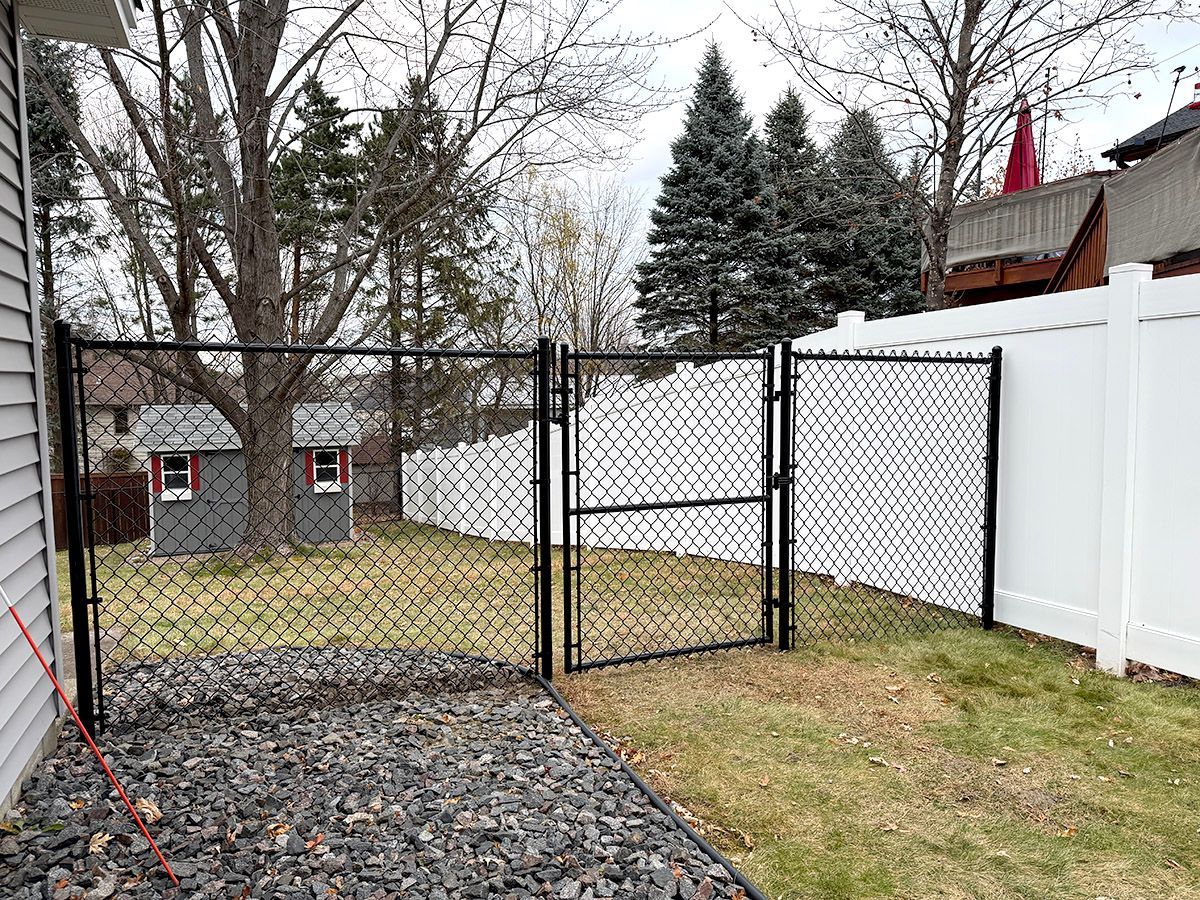 A black chain-link gate set between a house and a white vinyl fence in a backyard with a small shed and trees.