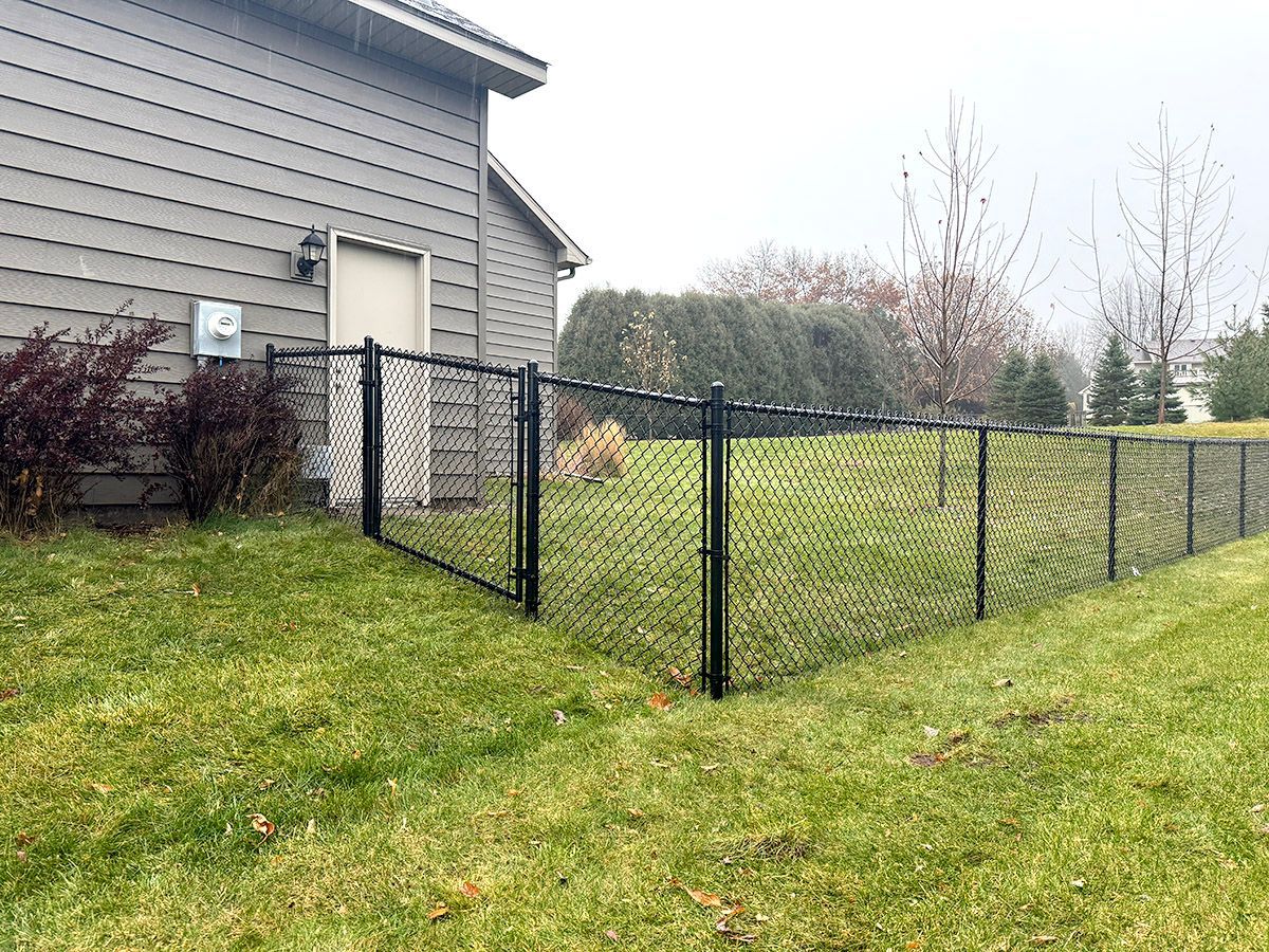 A black chain-link fence encloses a grassy backyard next to the side of a gray-sided house on an overcast day.