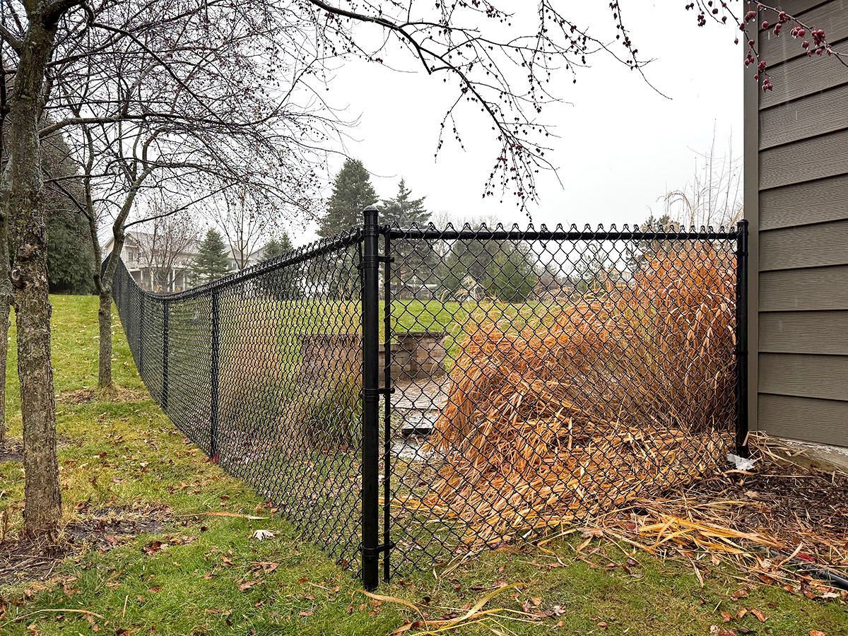 Black chain-link fence extending from a house exterior wall across a green lawn with trees and bushes in the background.