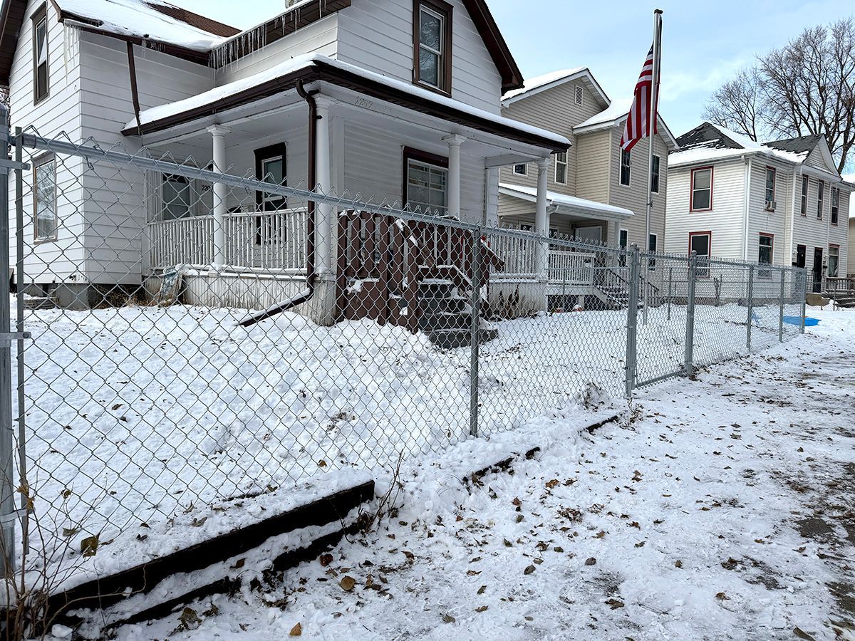 A snowy residential street featuring a white house with a front porch, enclosed by a chain-link fence.