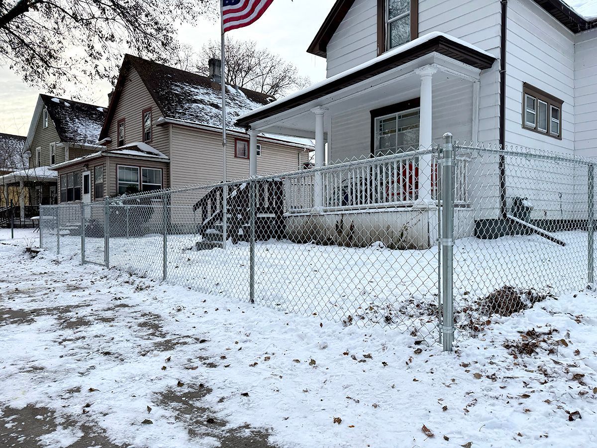 A snowy residential street featuring two houses behind a metal chain-link fence, with an American flag flying above.
