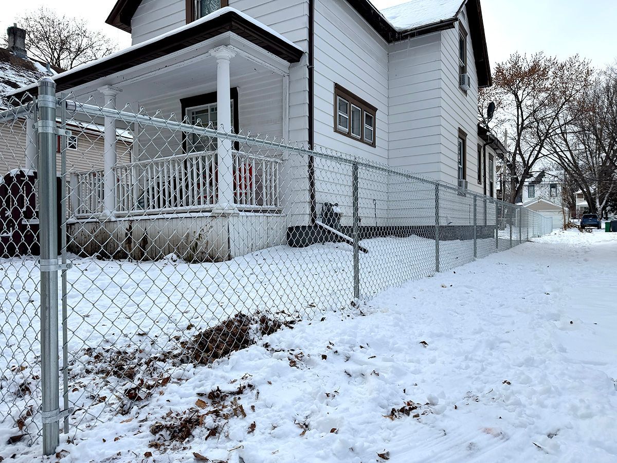 A white house with a porch stands behind a new chain-link fence on a snowy day.