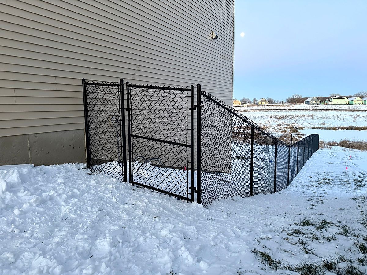 A black chain-link fence with a closed gate attached to the side of a beige building during winter with snow on the ground.