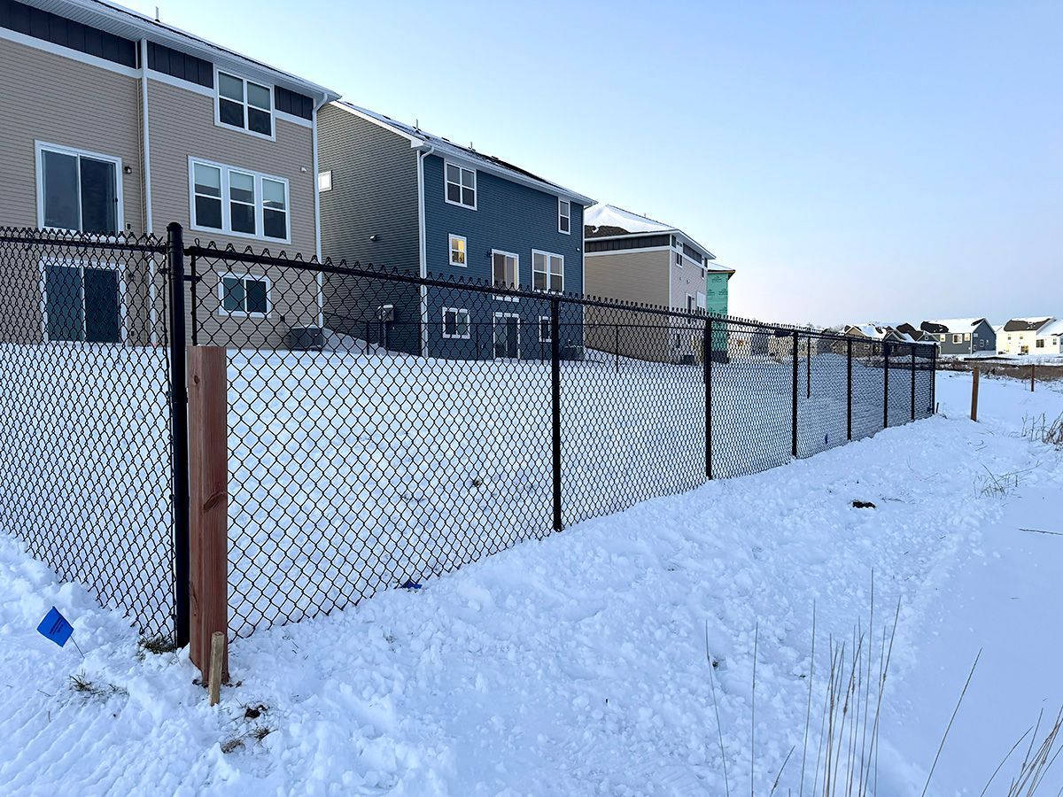 A black chain-link fence separates a snow-covered yard from several modern townhouses under a clear blue sky.