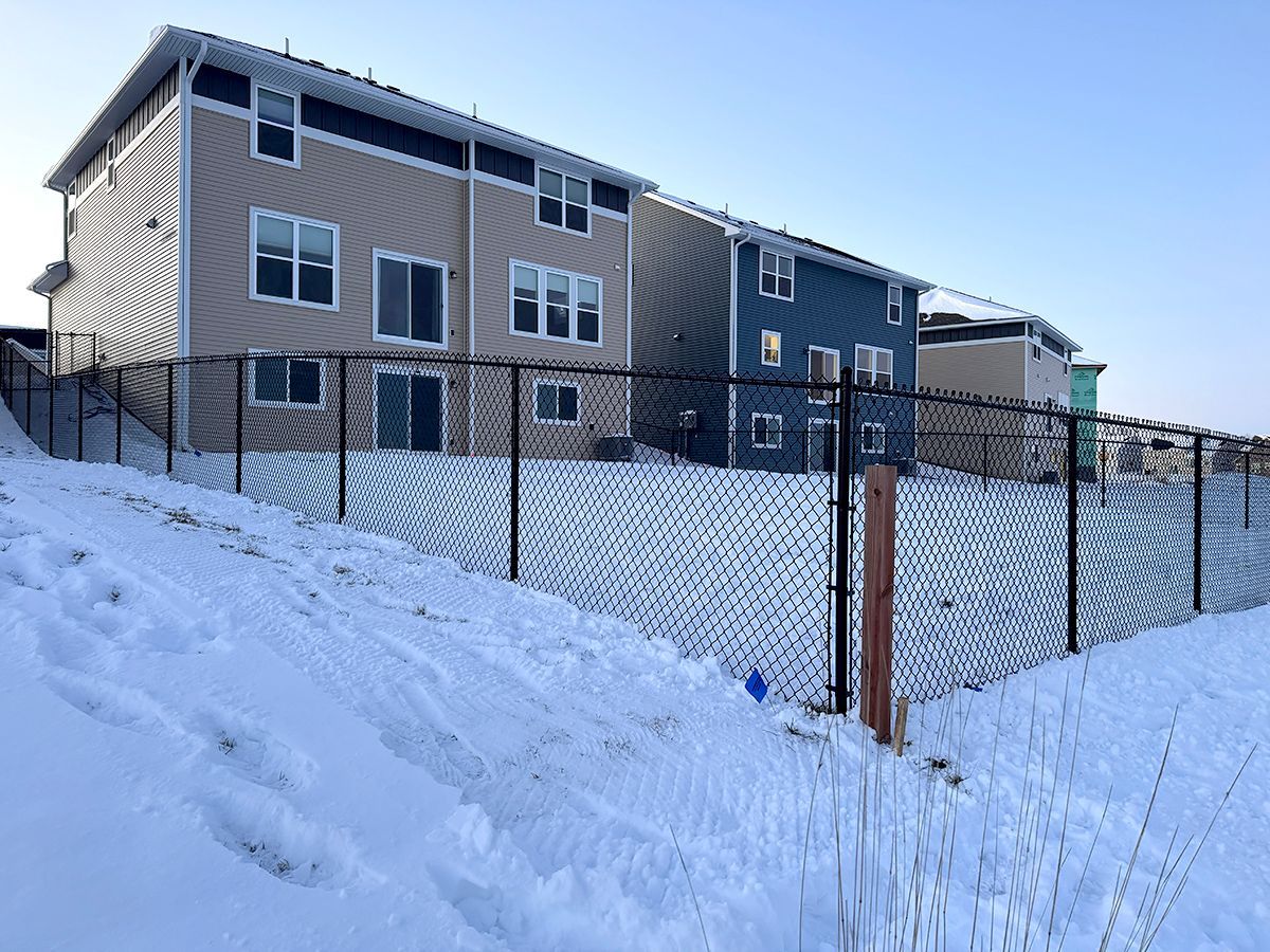 A snowy, sloped foreground with a chain-link fence separating two modern houses, one tan and one blue, against a clear sky.
