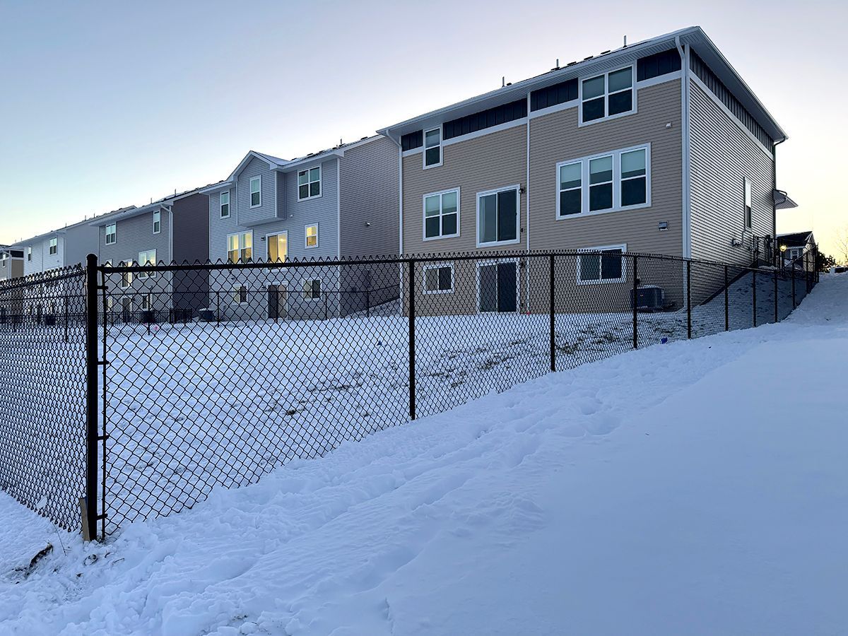A chain-link fence borders a snow-covered yard in front of several multi-story apartment buildings at dusk.