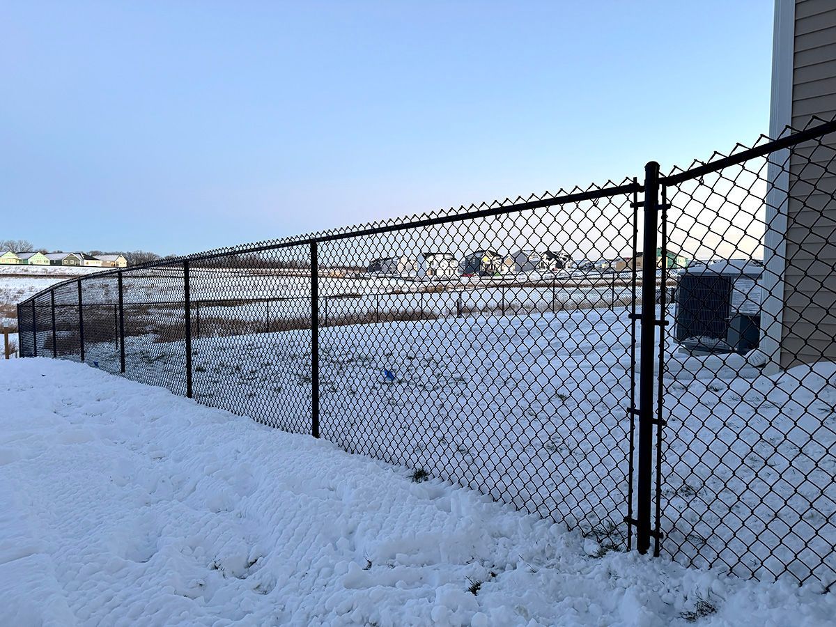 A black chain-link fence bordering a snow-covered backyard next to a house with a clear blue sky.