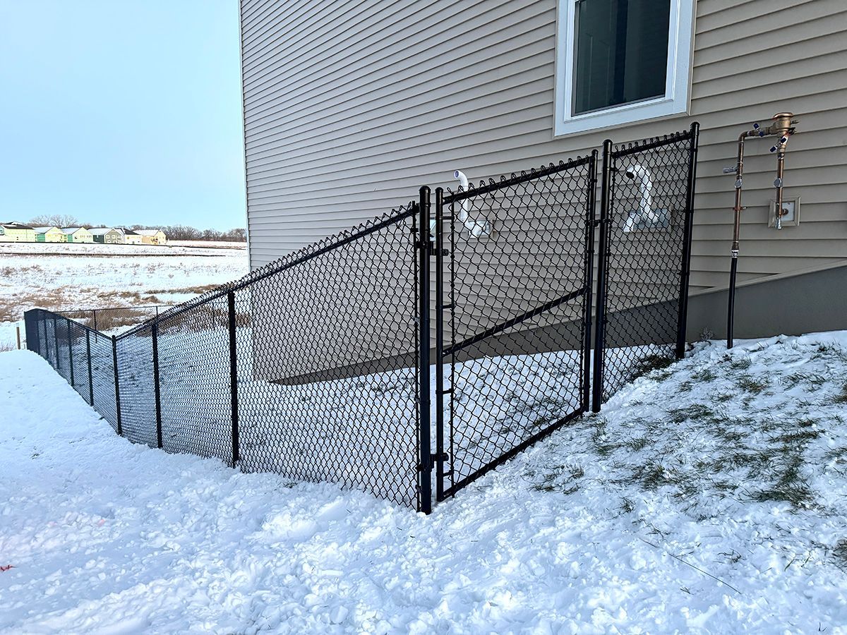 A black chain-link fence and gate installed on a snowy, sloped yard next to a tan house with siding.