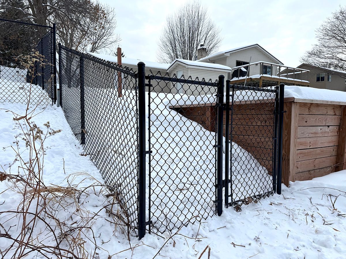 A black chain-link fence and a small gate enclose a snow-covered yard next to a wooden retaining wall in winter.