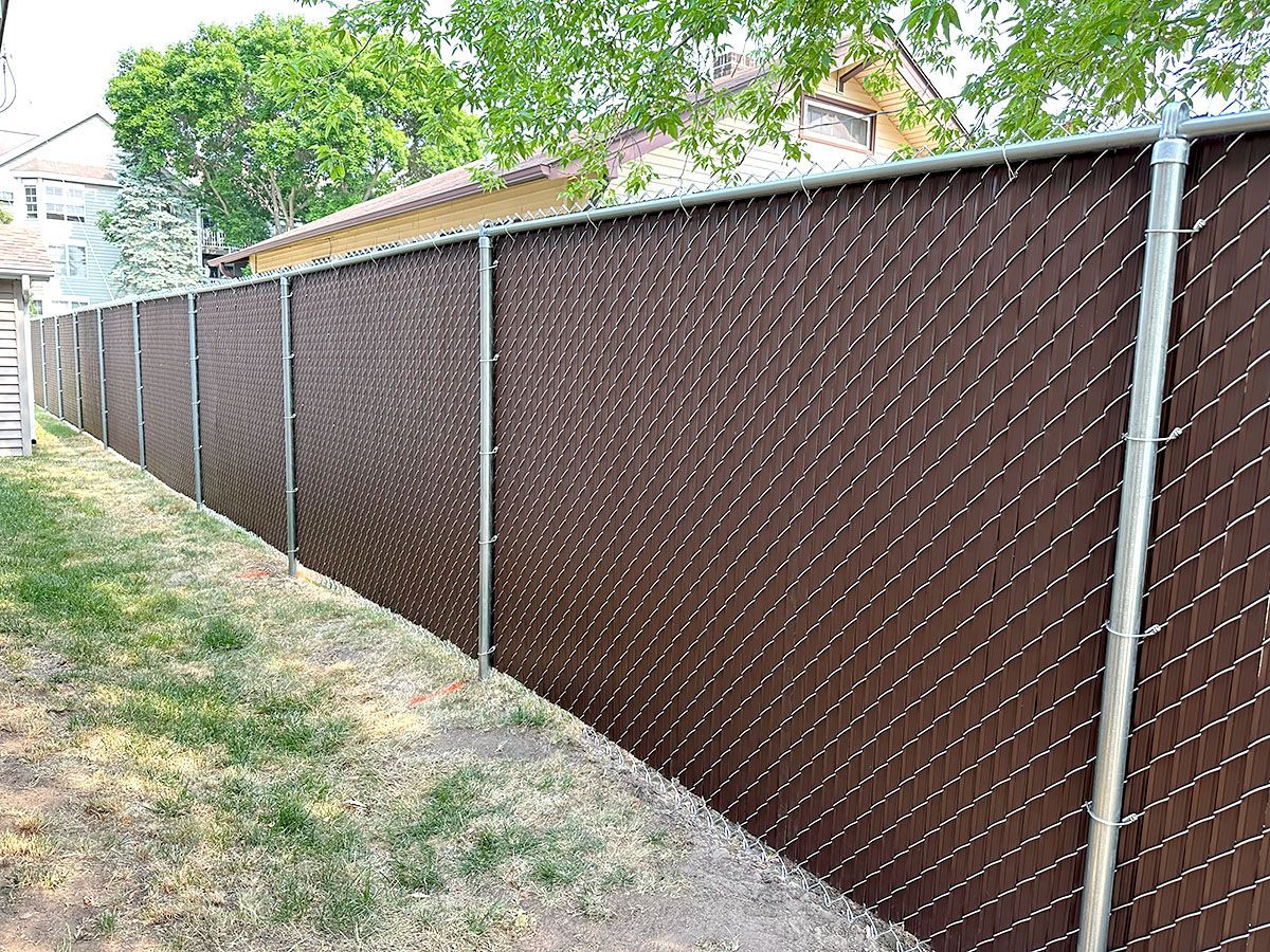 A brown, privacy-slatted chain-link fence standing along a grassy yard with houses in the background.