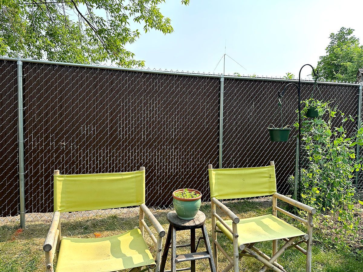 Two yellow canvas folding chairs sit on a patch of grass in front of a tall, brown chain-link fence with privacy slats.