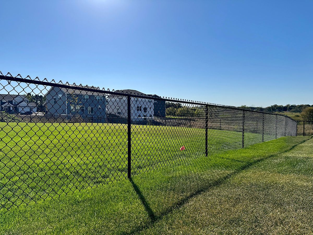 A black chain-link fence stretches across a green grass field under a clear blue sky, with houses in the background.