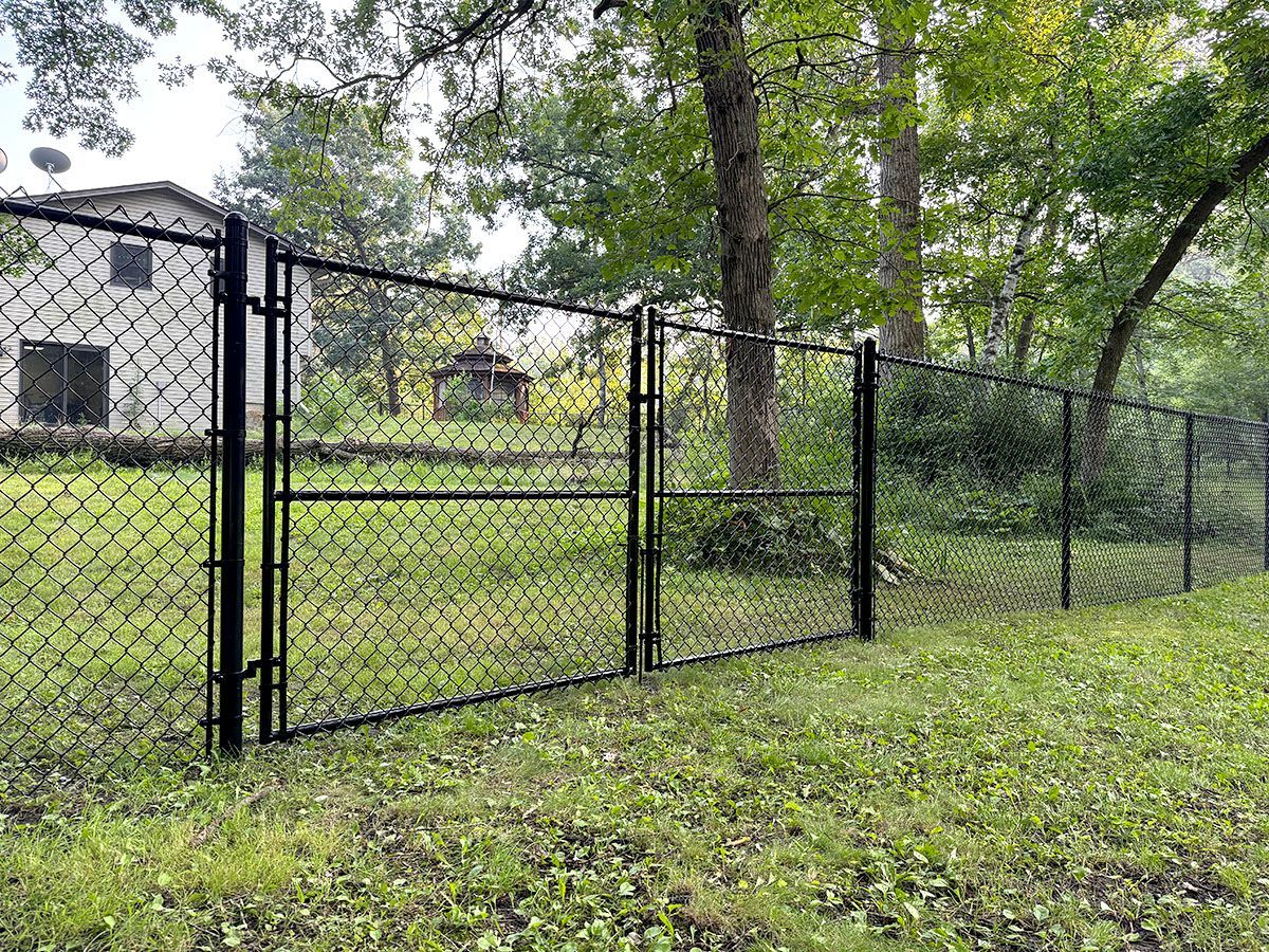 A black chain-link fence with a closed gate runs across a grassy backyard toward trees and a house in the background.