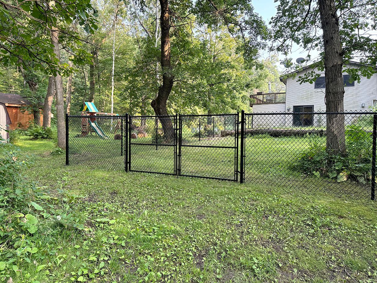 A black chain-link fence with a double-gate entrance spans a green, grassy backyard with trees and a house in the distance.