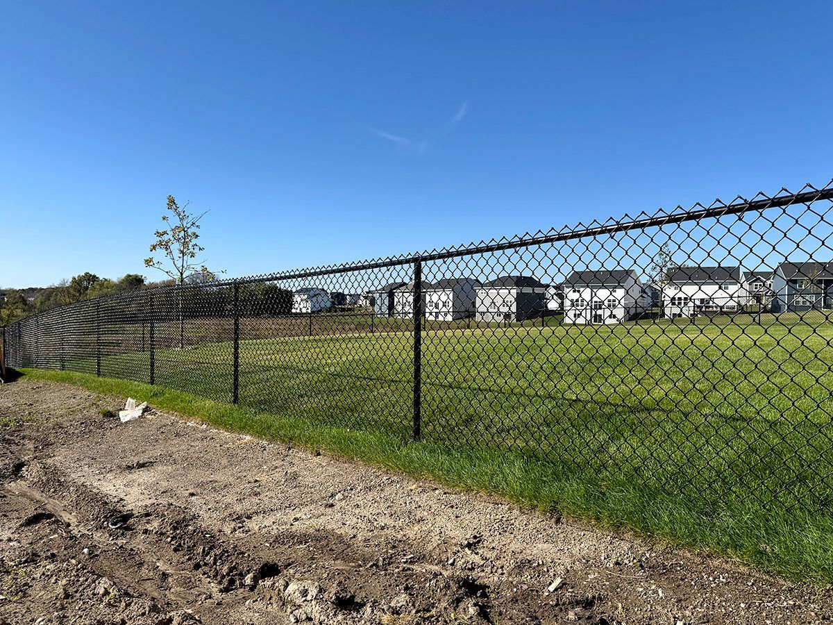 A black chain-link fence encloses a green grassy lot in front of suburban houses under a clear blue sky.