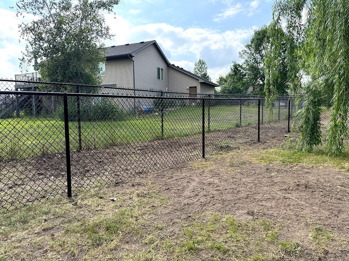 A black chain-link fence runs through a grassy yard toward a two-story beige house under a blue, cloudy sky.