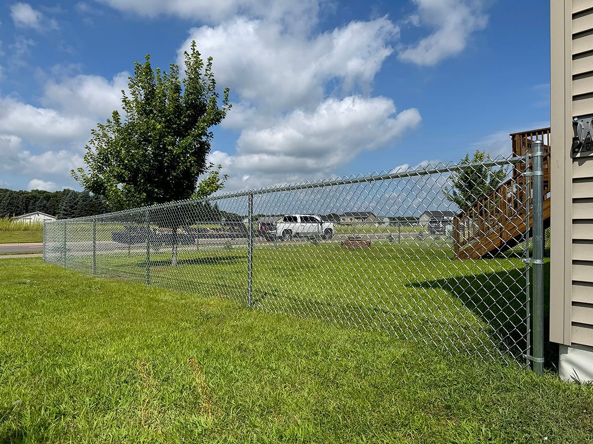 A chain-link fence encloses a grassy backyard on a sunny day, with a wooden deck and a tree visible nearby.