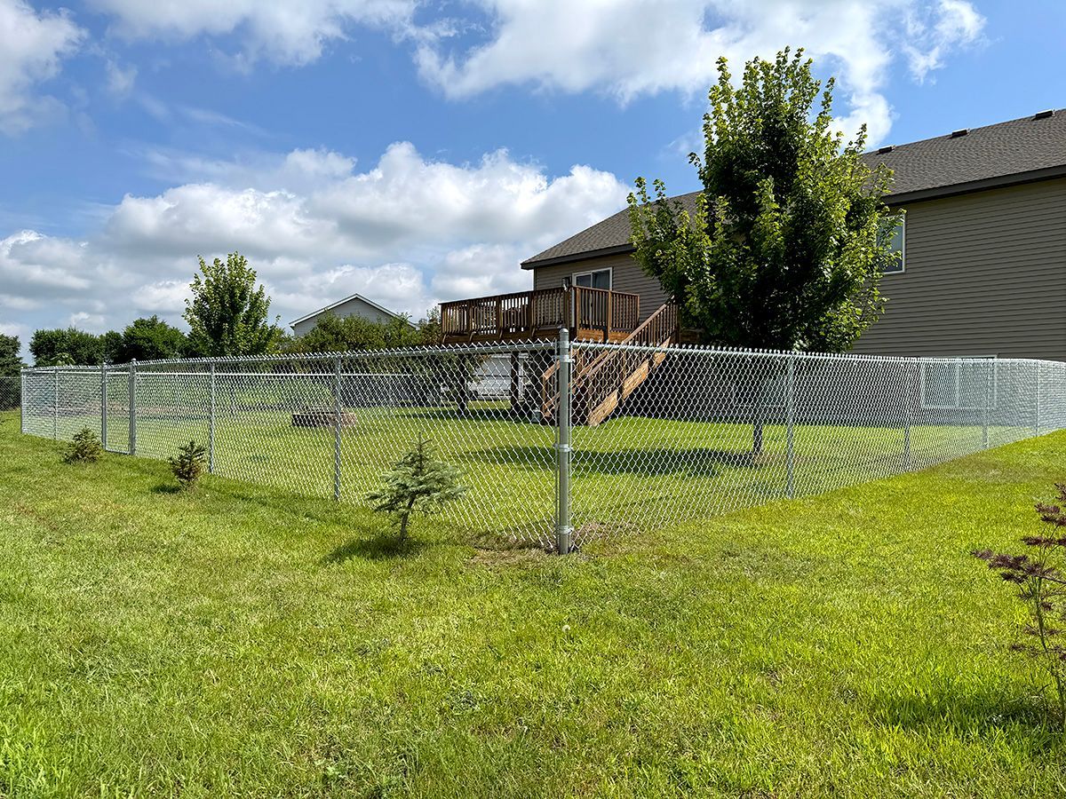 A chain-link fence surrounds a grassy backyard with a wooden deck and a house under a cloudy blue sky.