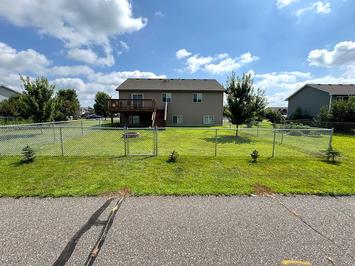 A two-story tan house with a wooden deck, surrounded by a chain-link fence in a suburban grassy yard under a cloudy sky.