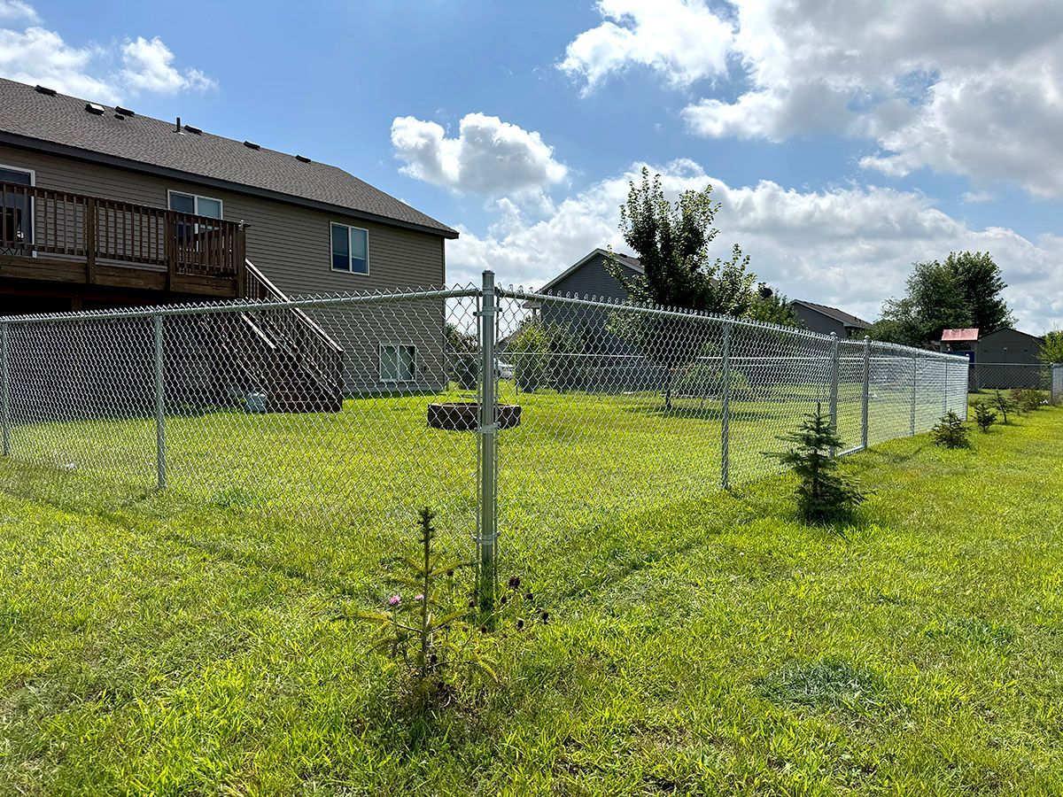 A fenced-in backyard with a brown house and wooden deck under a blue sky with white clouds.