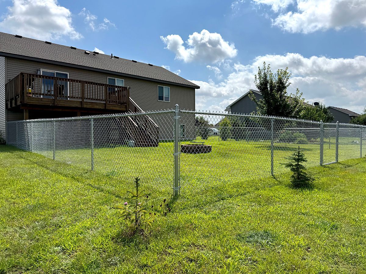 A two-story tan house with a wooden deck stands behind a chain-link fence in a grassy backyard under a partly cloudy sky.