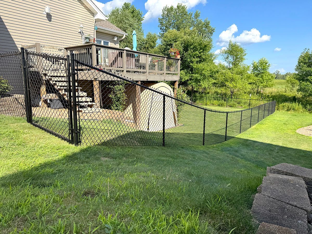 A black chain-link fence encloses a green backyard, extending from a house deck with stairs to a small storage shed.