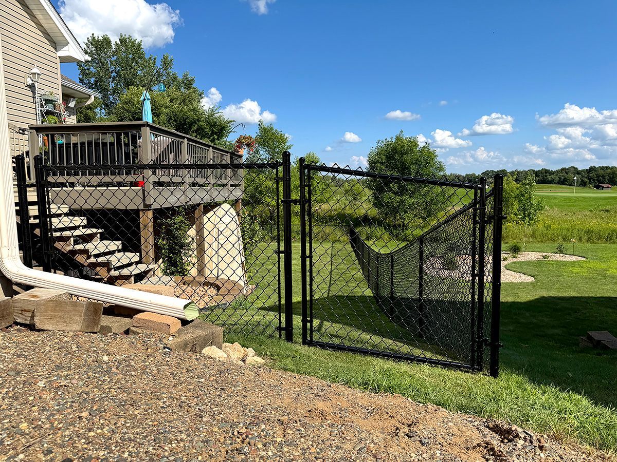 A black chain-link fence gate is open in a grassy backyard, next to a deck attached to a house.