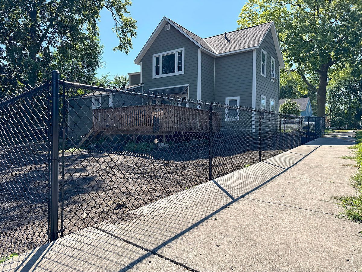 A two-story grey house with a wooden deck, enclosed by a black chain-link fence along a sidewalk on a sunny day.