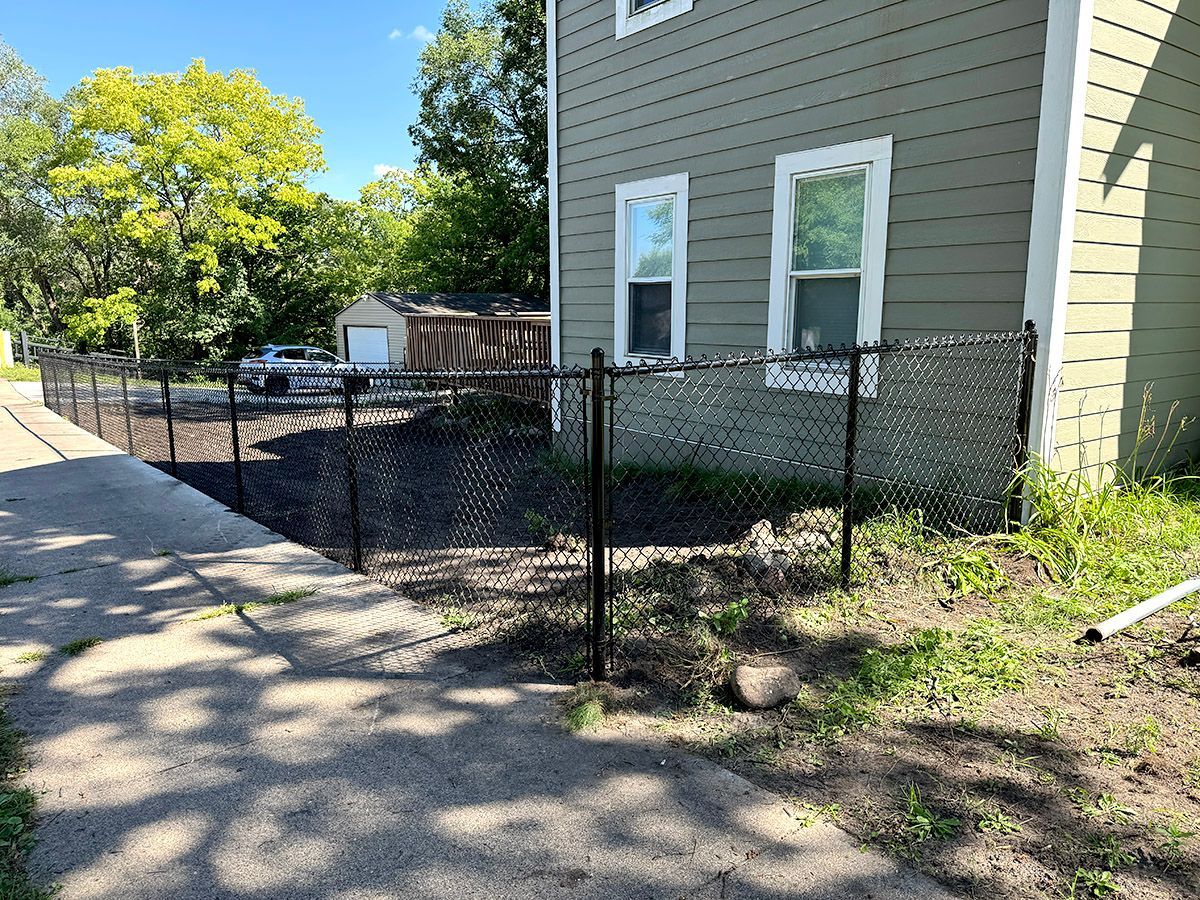 A black chain-link fence runs along the side of a light green house next to a paved sidewalk on a sunny day.