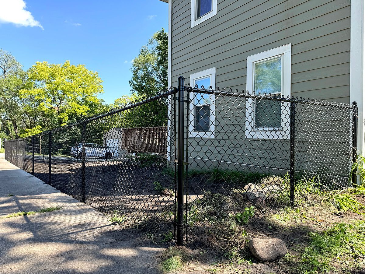 A black chain-link fence runs along a sidewalk beside a light green house on a sunny day.