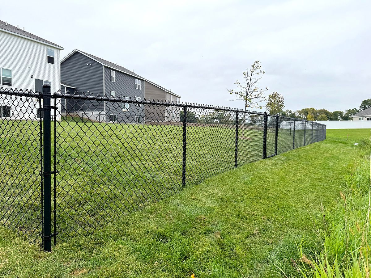 A black chain-link fence stretches across a grassy residential backyard beside two suburban houses.