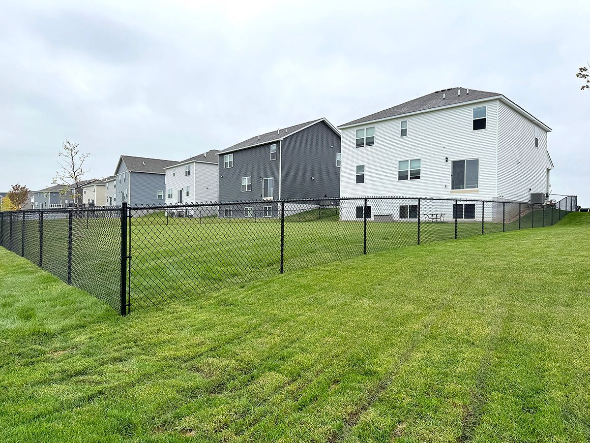 A black chain-link fence encloses a green grassy backyard behind a row of suburban houses on a cloudy day.