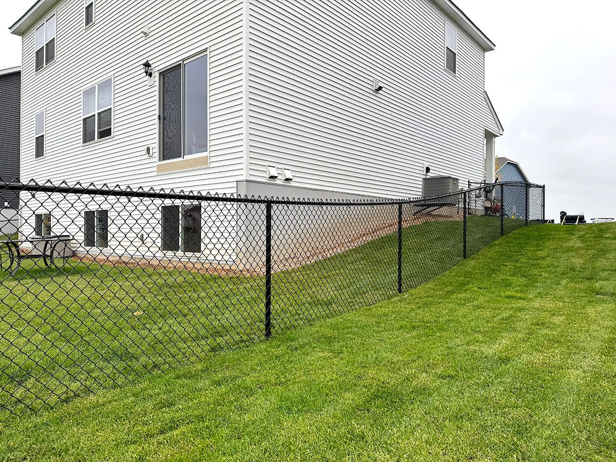 A white house with a black chain-link fence on a grassy, sloped residential yard under an overcast sky.